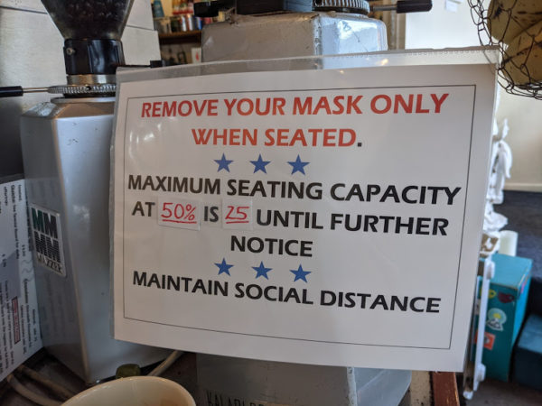 A sign saying 'remove your mask only when seated, maximum seating capacity at 50% is 25 until further notice" taped onto a coffee bean grinding machine