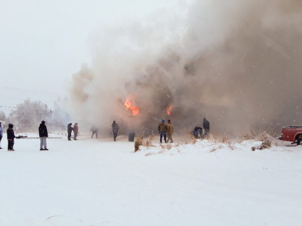 People gather in a snowy field as a smoke rises from a building