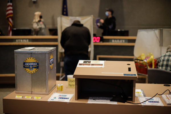 a ballot drop box sits on a table next to a podium. two people are in voting booths. two people are standing in the background