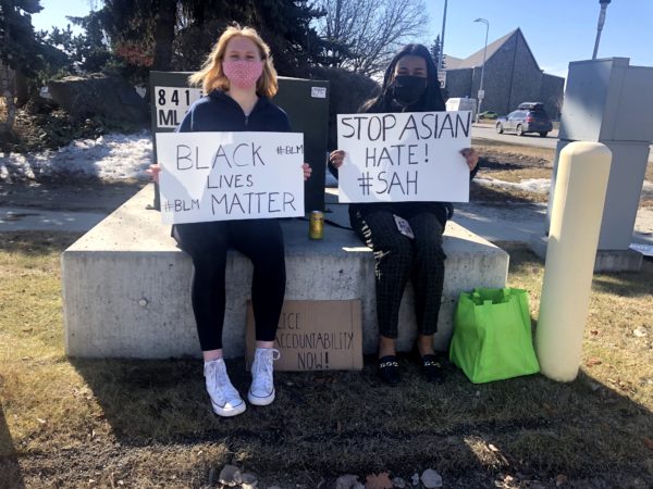 Two girls sit on a utility box on a street corner holding signs that read "Stop Asian Hate" and "Black Lives Matter"