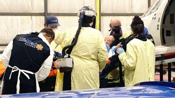 A woman surrounded by medical professionals inn PPE on a stretcher by a plane
