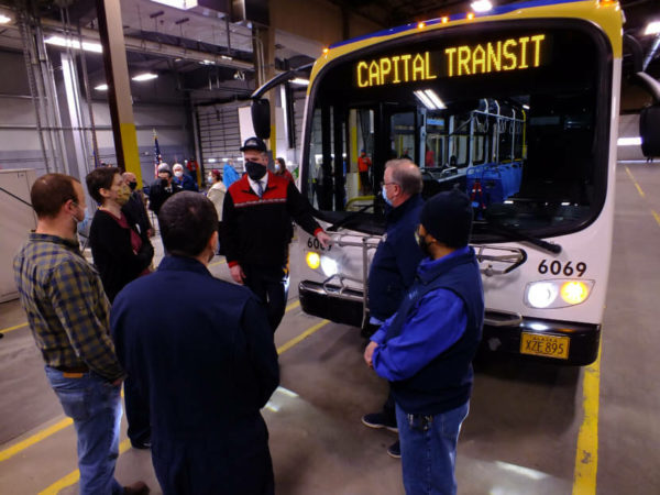 A crowd gathers around the front of a bus in a warehouse