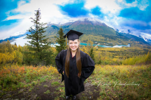 A high school senior in a cap and gown poseses in front of a green mountain