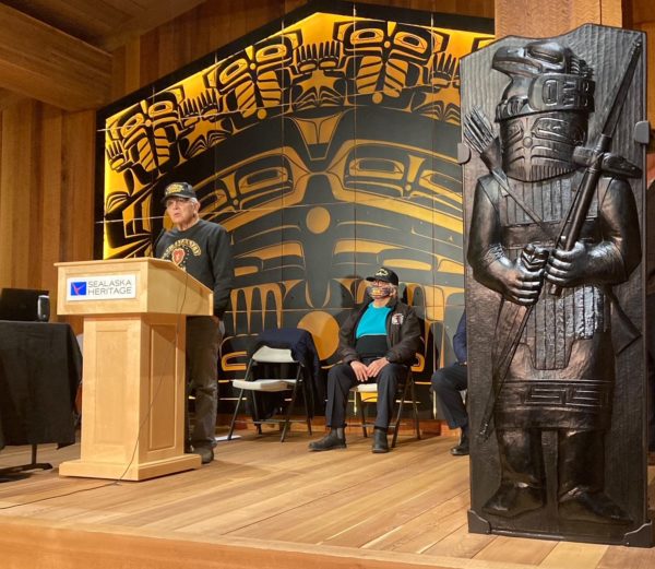 An Alaska Native person speaks at a podium in front of Tlingit motifs