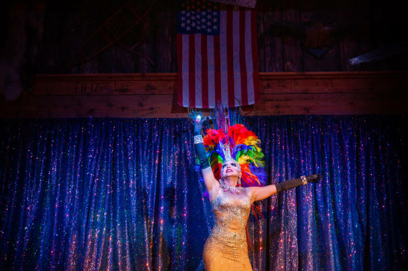 A person in drag stands on stage raising their hands