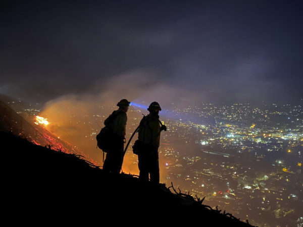 Shadows of two firefighters are backlit by flames, overlooking a city at night.