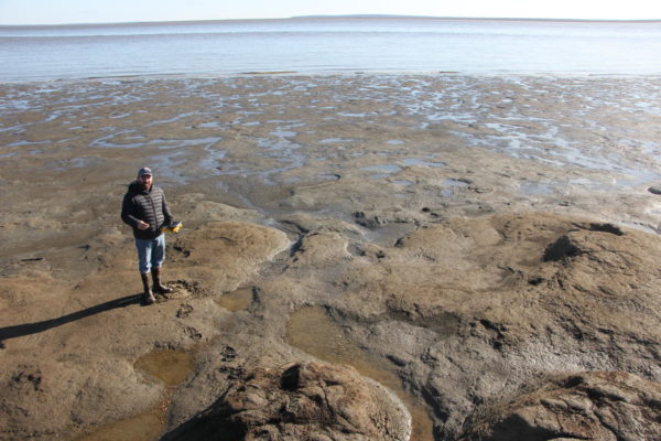 A man in a winter jacket and boots stands on a muddy patch of a swampy lagoon.