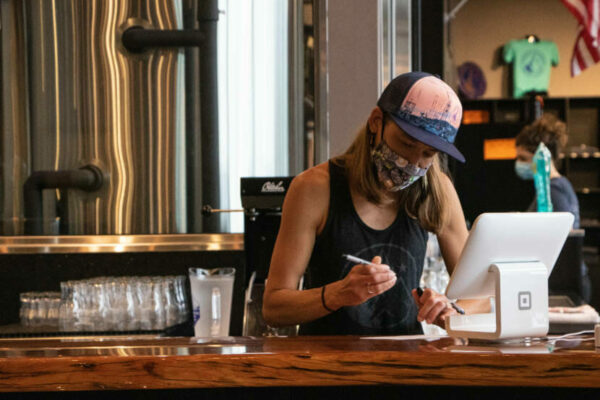 A woman ina trucker hat writes on a paper pad in front of a brewing vat