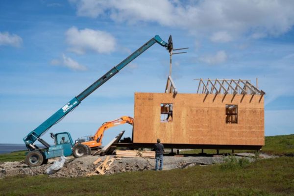 A crane and backhoe put up a house in a green field
