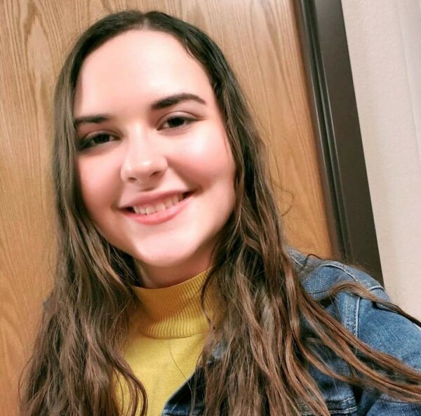 A young woman poses for a selfie in a yellow turtleneck and jean jacket.