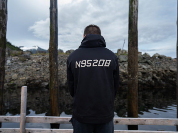 A man with his back turned stands in a dock facing the water. The back of his black sweathshirt has numbers 