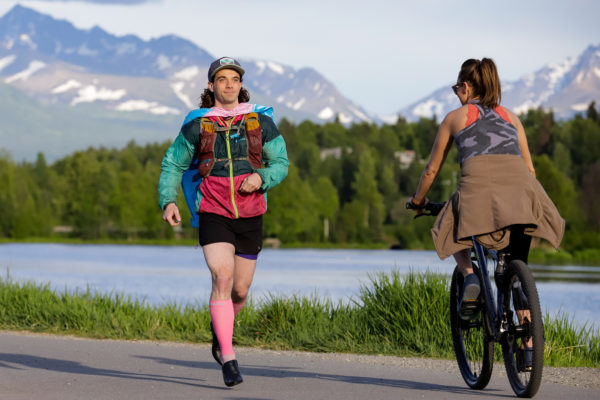 a person wearing sportswear, high heels, and a cape jogs by a biker in front of a body of water