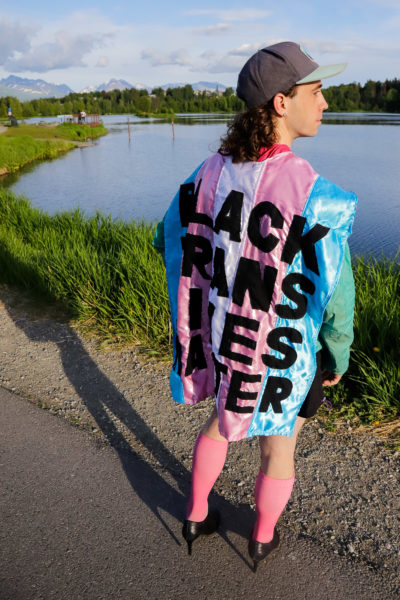 a person wearing sportswear, high heels, and a cape that reads "BLACK TRANS LIVES MATTER" poses in front of a body of water