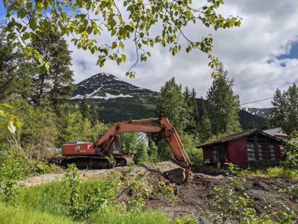 A backhoe digs a hole next to a red little house in front of a mountain