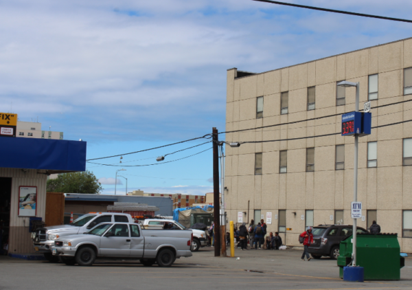 A gas station garage next to a parking lot in front of a tan building where people are gathered