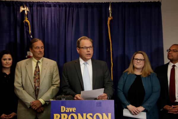 A white man speaks at a podium in front of a blue "Dave Bronson sign" On his left is a white man with a tan suit, on his right is a white woman with a blue jacket and blond hair