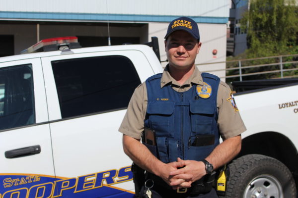 A white man wearing a state trooper uniform stands in front of a white truck with his hands clasped in front of him.