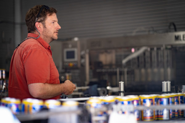 A man stands behind a line of beer cans moving down a conveyer belt.