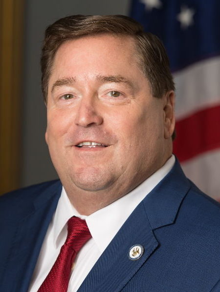 A white  man with brown hair a blue suit and red tie poses in front of the American flag