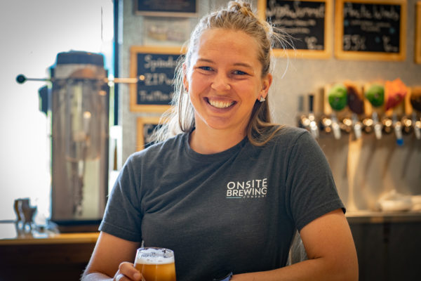 A woman stands in front of beer taps, holding a glass of beer in her hand.