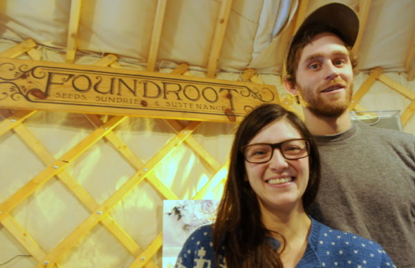 A man and woman stand inside of a yurt, in front of a wooden sign that says Foundroot.