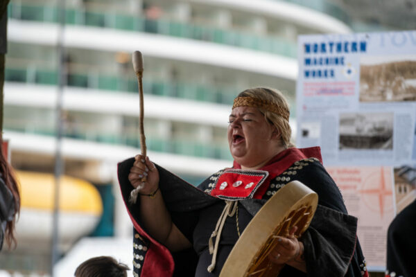 A woman in a black Tlingit-patterned robe beats on a drum and sings
