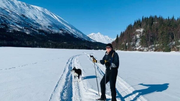 A man stands in the snow with ski poles, his dog nearby.
