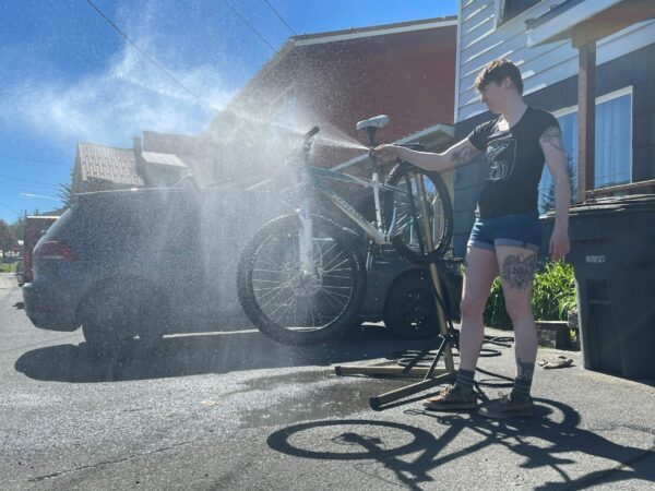 A woman in shorts and a t-shirt sprays down a mountain bike with a hose.