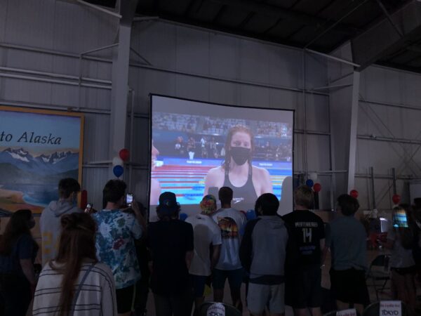 A group of people stand in a big room watching a screen with a swimmer's image projected on it.