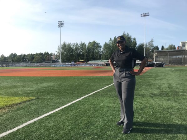 A woman in a baseball umpire's uniform stands with her hands on her hips on the third baseline of a baseball diamond.