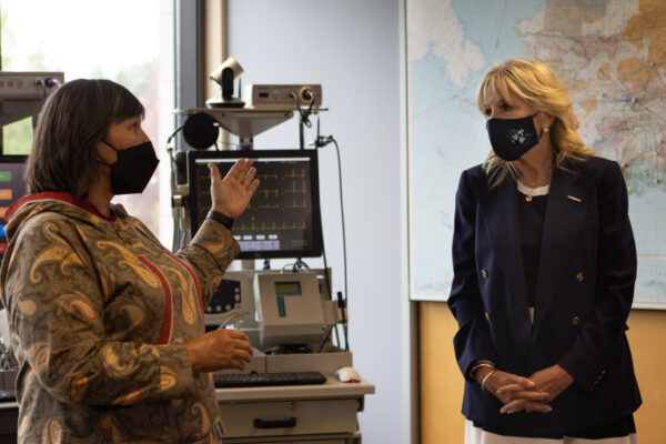 Two women talk, both wearing face masks, near medical equipment.