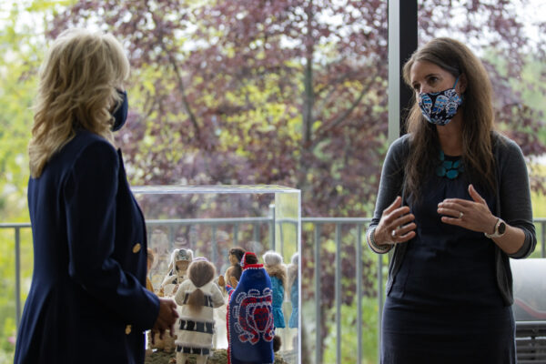 Two women wearing face masks talk inside, near big windows.