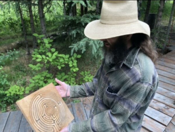 A man in a flannel shirt, a white straw hat holds a board of wood with a circular labyrinth design with some forest in the background