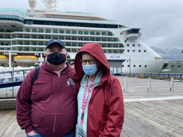 Two people stand in zipper-up jackets and sweatshirts in front of a cruise ship.