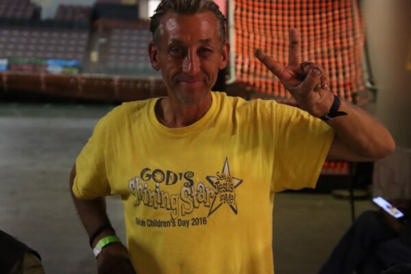 A man in a yellow t-shirt waves a peace sign
