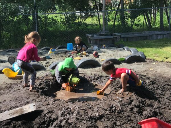 Children play in the mud and in a sandbox with trucks and other toys.
