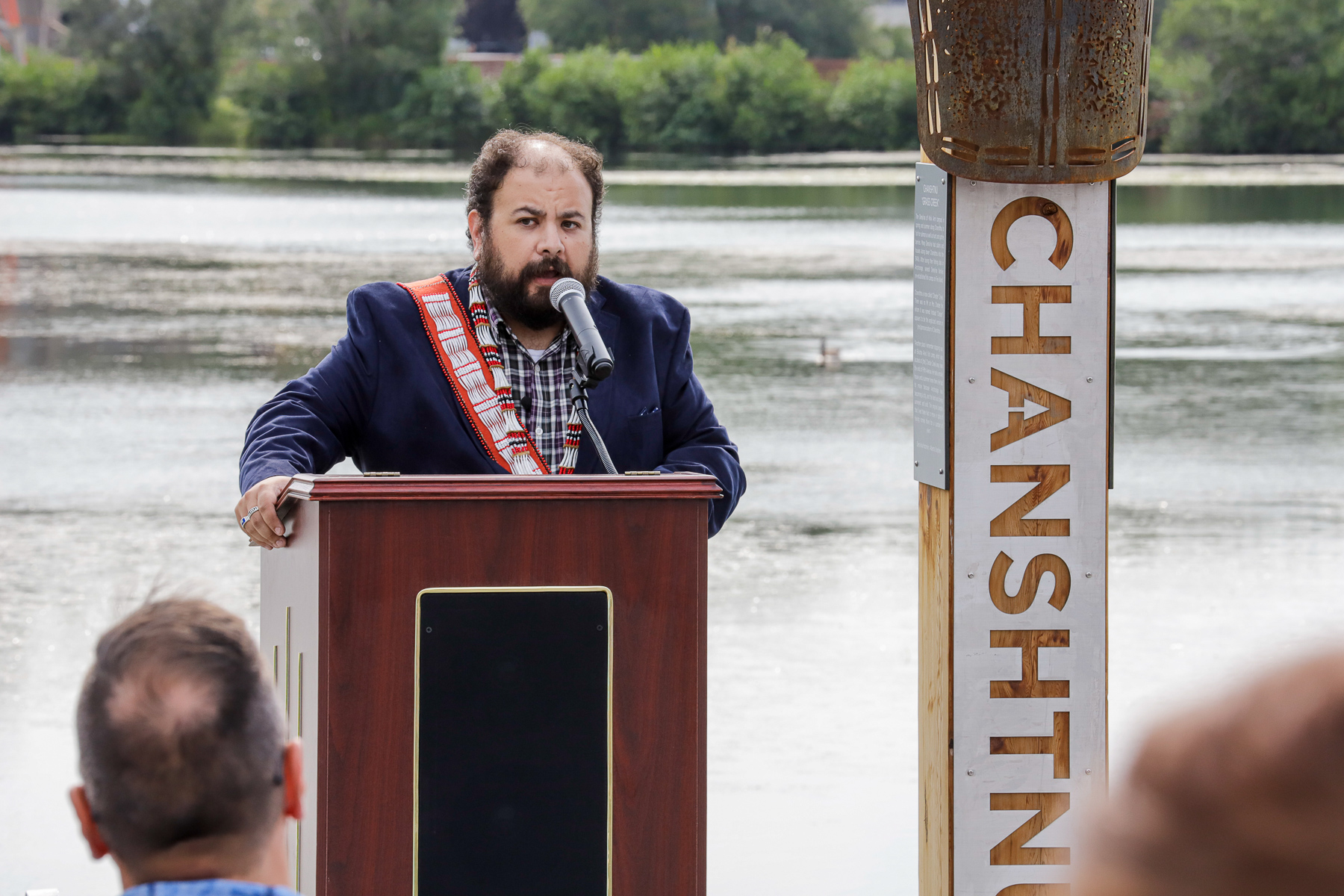 a person speaks from behind a podium, next to an art installation, in front of a lagoon
