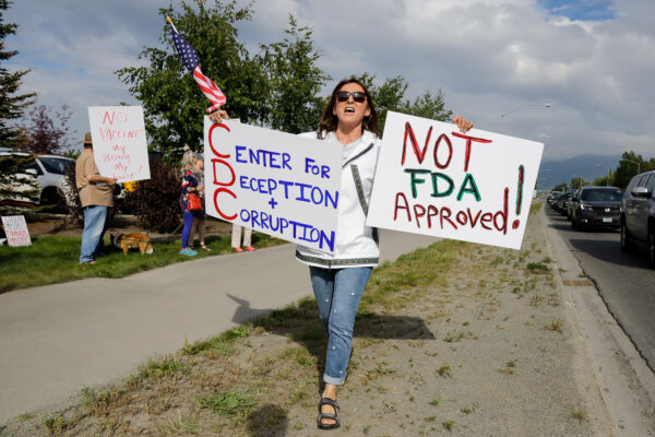 A woman in a white qaspek and jeans walks down the grass strip next to a bus road holding a sign that says "Not FDA approved" and Center for Deception and Corruption