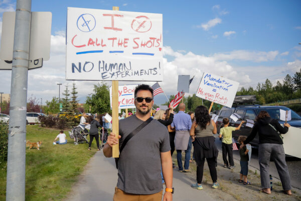 A white man in a tshirt and tan pants holds a sign that says "I call the shots" in front of other protestors