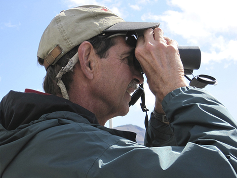 A man in a cap and jacket looks through binoculars.