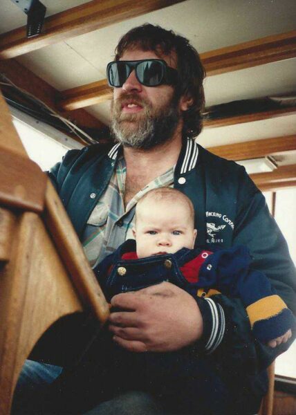 A baby sits on a man's lap who is wearing sunglasses and steering a boat.