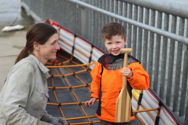 A woman smile as she looks at a toddler who is holding a small wooden canoe paddle.
