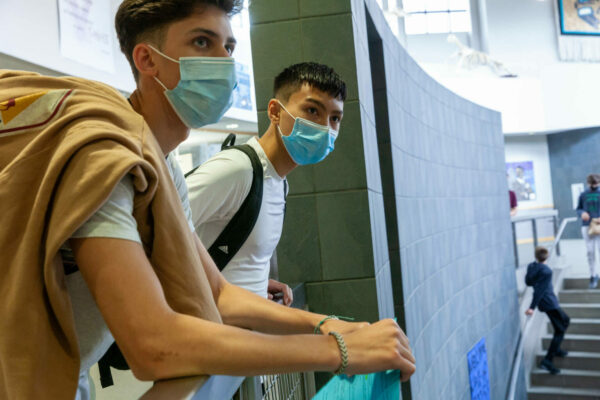 Two teenagers in face masks look over a stairs at school.