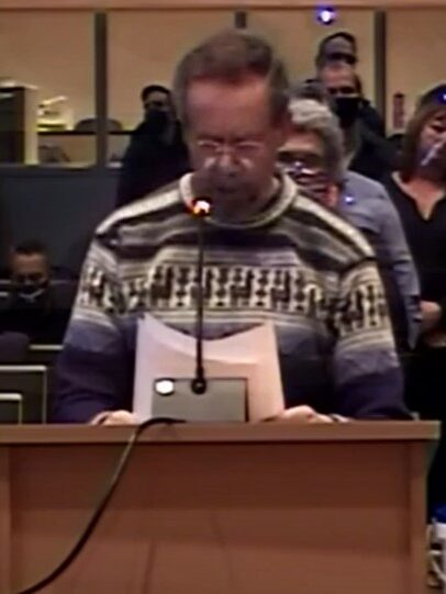 A white man in a patterned wool sweater speaks into a microphone at a podium