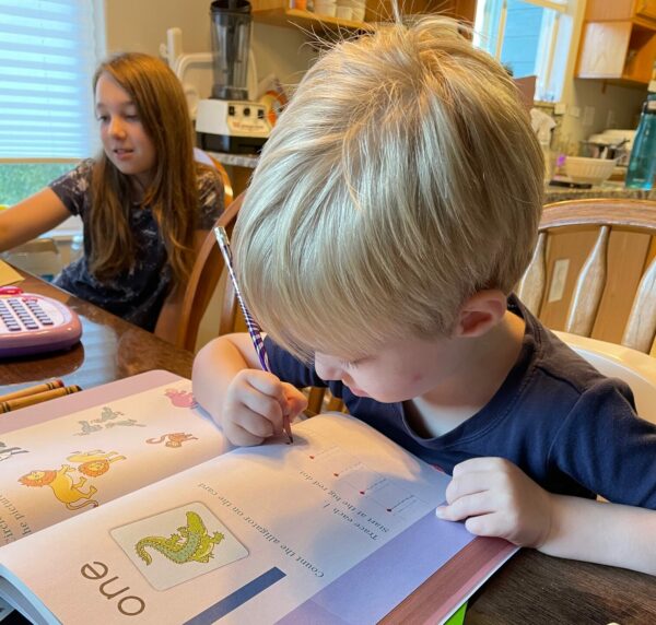 A toddler boy with blonde hair works in a workbook at a table next to an older girl with brown hair also doing school work at the table.