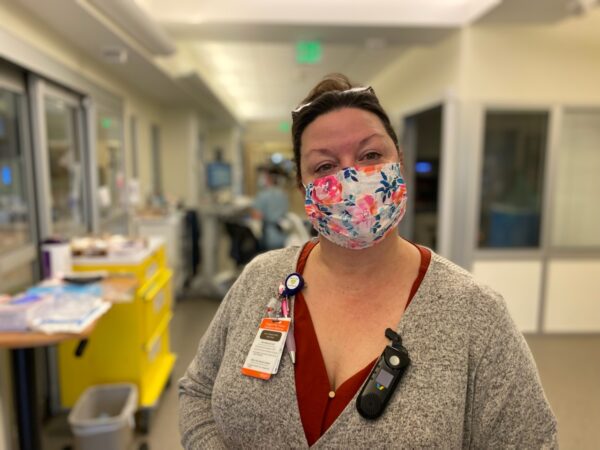 A woman in a flowered mask and a greey sweater smilees in a medical hallway