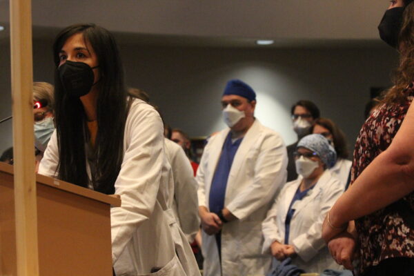 woman in lab coat and mask stands at podium with others behind her.
