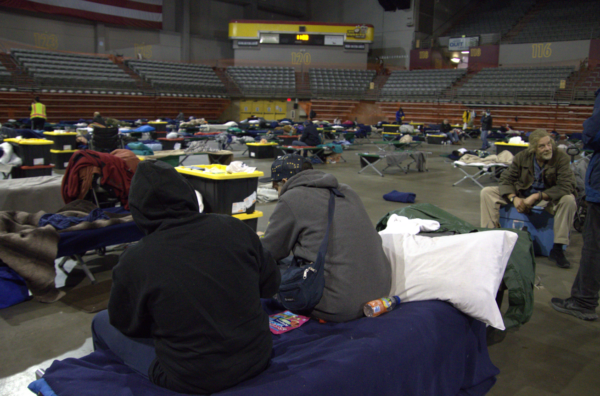People sit on their cots on the floor of an arena