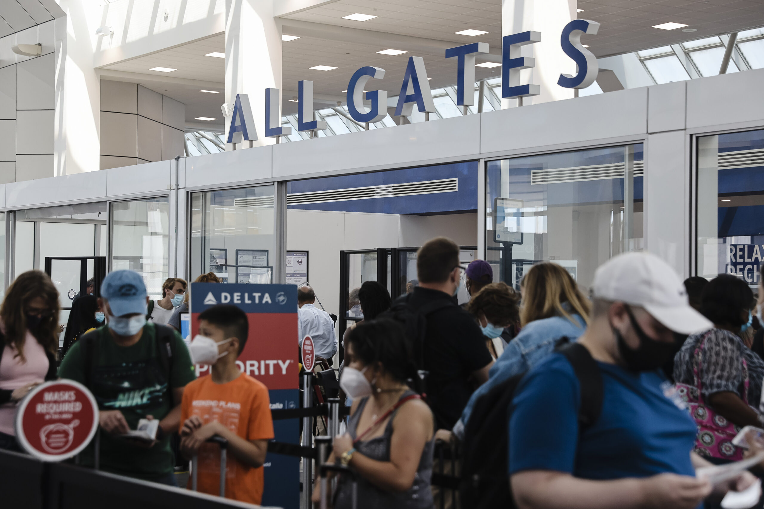 People stand in line around a sign that says "All Gates" wearing masks.