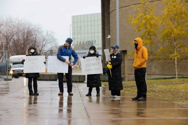 Five people stand outside with masks on and in winter coats holding signs.